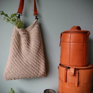 May include: A beige crocheted tote bag with a brown leather strap hanging on a wall. Two brown leather cylindrical bags are stacked on a table below.