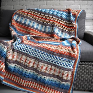 Puede incluir: Una manta de ganchillo de colores con rayas azules, naranjas, marrones y blancas. La manta está colocada sobre un sofá de mimbre gris.