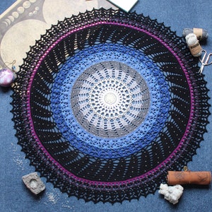 Op de afbeelding: Een groot, rond, zwart gehaakt doily met een blauw, grijs en wit centrum. Het doily heeft een gegolfde rand en is versierd met kleine kralen.