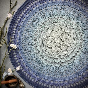 Puede incluir: Un gran mandala redondo tejido a crochet en tonos azules y blancos. El mandala tiene un diseño delicado e intrincado y está hecho con un hilo degradado que pasa del azul oscuro al azul claro al blanco.