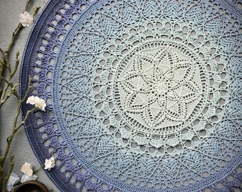 Puede incluir: Un gran mandala redondo tejido a crochet en tonos azules y blancos. El mandala tiene un diseño delicado e intrincado y está hecho con un hilo degradado que pasa del azul oscuro al azul claro al blanco.