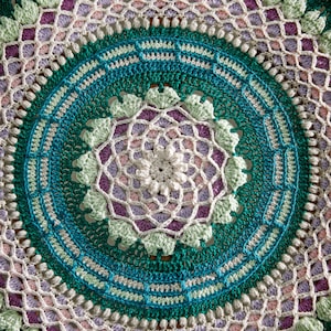 May include: A large, round crocheted mandala with a teal, green, white, pink, and purple color scheme. The mandala is made with a variety of stitches and has a detailed, intricate design.