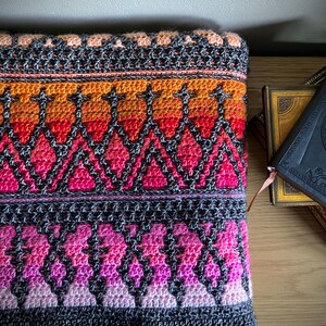 Op de afbeelding: Een kleurrijke, handgehaakte deken met een geometrisch patroon. De deken heeft een kleurengradatie van oranje, rood, roze en paars, met een donkergrijze rand. De deken is opgevouwen en ligt op een houten oppervlak.