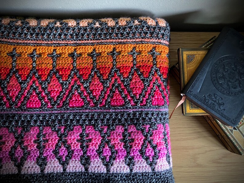 Op de afbeelding: Een kleurrijke, handgehaakte deken met een geometrisch patroon. De deken heeft een kleurengradatie van oranje, rood, roze en paars, met een donkergrijze rand. De deken is opgevouwen en ligt op een houten oppervlak.