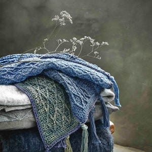 Może przedstawiać: Stosik dzianych kocy w odcieniach niebieskiego i zielonego z frędzlami. Koce są ułożone na drewnianym stołku z gałązką suszonych kwiatów.