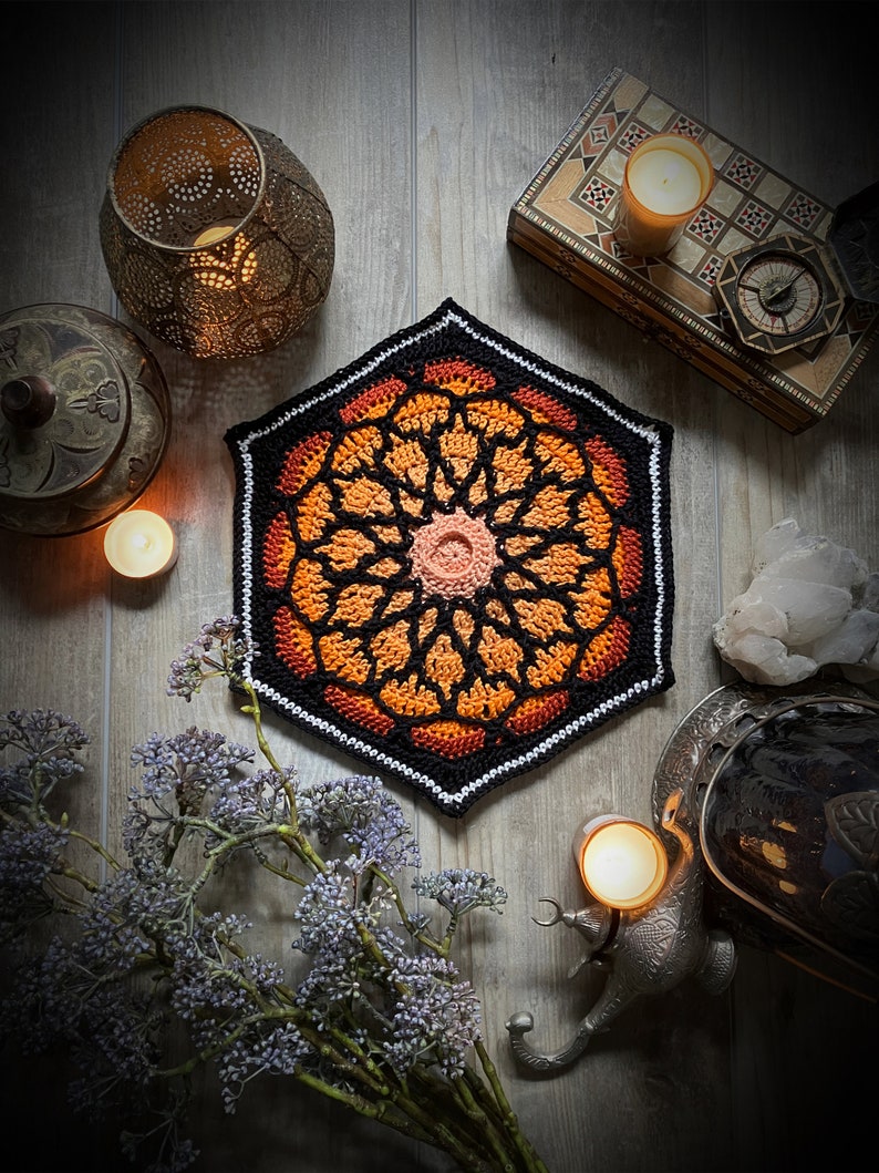May include: A crocheted hexagonal mandala with a black, orange, and white color scheme. The mandala is surrounded by candles, lanterns, and other decorative items.