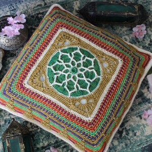 Puede incluir: Una almohada de crochet con un diseño de mandala verde y blanco en el centro. La almohada está rodeada por un borde multicolor con detalles dorados.
