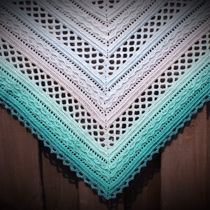 May include: A handmade, triangular crochet shawl in shades of teal, blue, and lavender. The shawl features a delicate openwork design with a scalloped edge. The gradient color scheme adds depth and visual interest to the soft, textured fabric.
