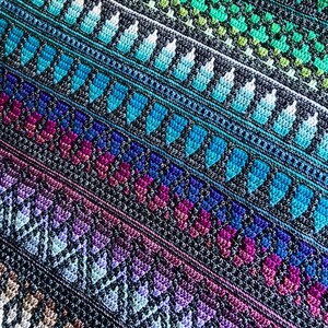 Op de afbeelding: Een close-up van een kleurrijke, handgemaakte gehaakte deken. De deken heeft diagonale strepen met verschillende patronen en kleuren, waaronder blauw, groen, paars en beige. Het ingewikkelde stekenwerk cre&euml;ert een textuurrijk en visueel aantrekkelijk ontwerp.