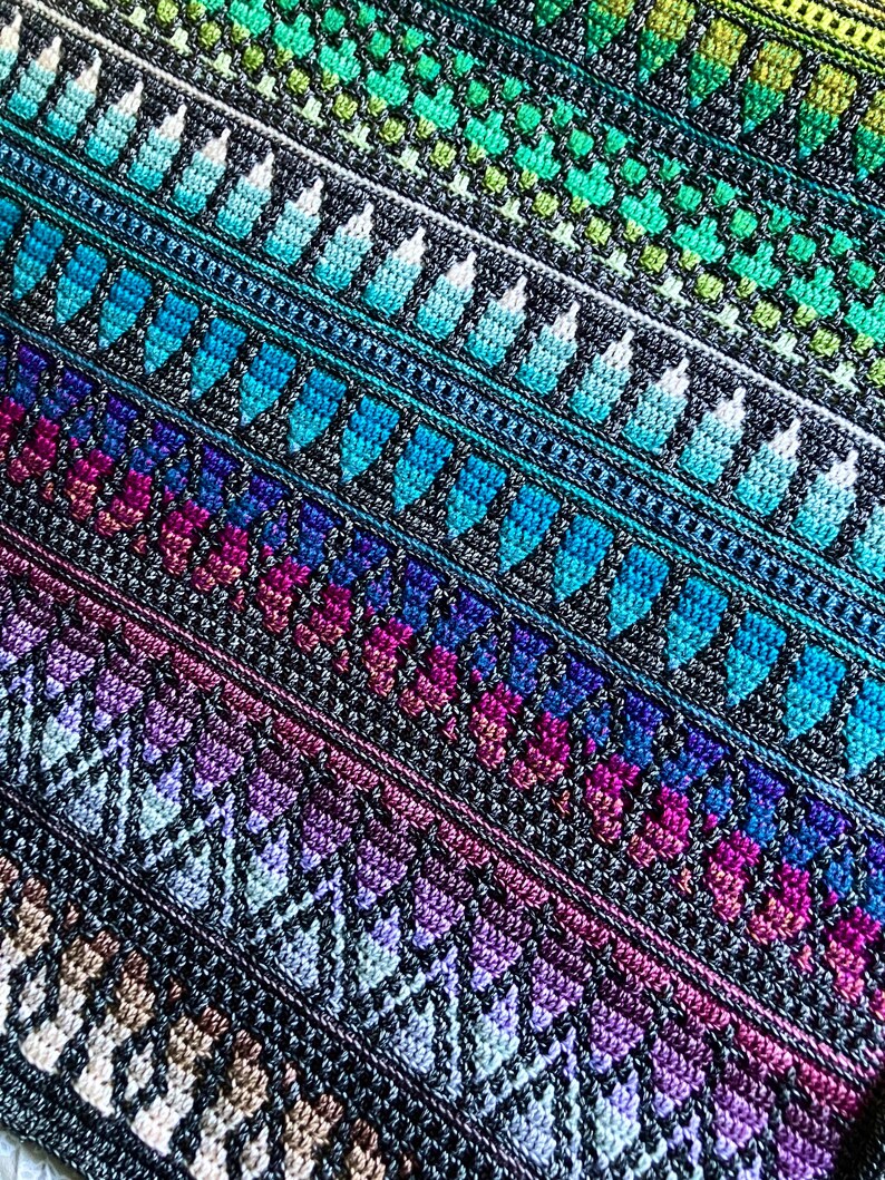 Op de afbeelding: Een close-up van een kleurrijke, handgemaakte gehaakte deken. De deken heeft diagonale strepen met verschillende patronen en kleuren, waaronder blauw, groen, paars en beige. Het ingewikkelde stekenwerk cre&euml;ert een textuurrijk en visueel aantrekkelijk ontwerp.