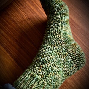 Op de afbeelding: Groene en gele gebreide sok met een textuurpatroon. De sok is gemaakt van garen in tinten groen, geel en bruin. De sok ligt op een houten oppervlak.