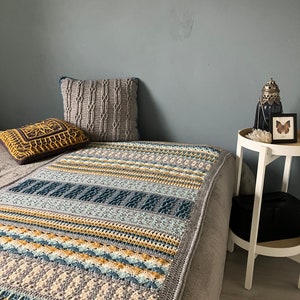 Könnte beinhalten: Eine graue und weiße gehäkelte Wolldecke mit gelben und blauen Streifen. Die Decke liegt auf einem Bett mit zwei Kissen, einem grauen und einem braunen mit einem gelben Muster.