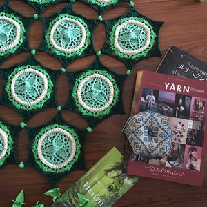 May include: A green and white crocheted blanket with a geometric pattern. The blanket is on a wooden surface with several origami cranes and books.