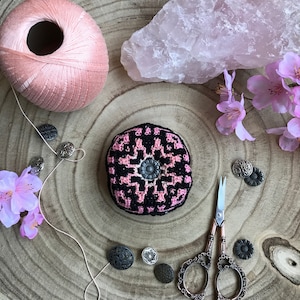 Puede incluir: Un alfiletero de crochet rosa y negro con un botón en el centro, rodeado de botones vintage, una bobina de hilo rosa, un cristal de cuarzo rosa y un par de tijeras antiguas sobre una superficie de madera.