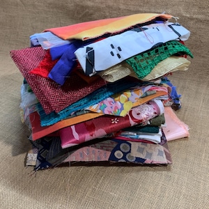 May include: A pile of colourful fabric scraps in various textures and patterns. The fabrics include red, blue, green, and floral prints. The scraps are stacked on a hessian surface.