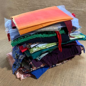 May include: A pile of assorted fabric remnants in a variety of colours and textures. The fabrics include orange, blue, green, and purple, with some patterned pieces. The stack rests on a textured, tan surface.