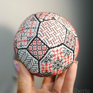 Op de afbeelding: Een witte bol met zwarte en rode borduurwerk, verdeeld in secties met verschillende patronen. De bol wordt in een hand gehouden.