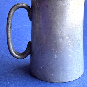 Puede incluir: Una jarra cilíndrica de peltre con un acabado gris mate y desgastado, que cuenta con un asa curva y lisa. La superficie del metal muestra sutiles signos de desgaste y textura, con un tono metálico consistente en todo el cuerpo del recipiente.