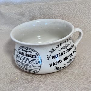 Könnte beinhalten: Weißer Keramiknapf mit Henkel, mit schwarzem Text und dekorativen Elementen. Der Napf hat einen schrägen Rand und einen gebogenen Henkel. Der Text enthält "Dandelion Cocoa" und "Patent Charcoal Rapid Water Filter".