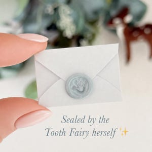 K&ouml;nnte beinhalten: Ein kleiner wei&szlig;er Briefumschlag mit einem hellblauen Wachssiegel, das einen Handabdruck zeigt. Unter dem Umschlag steht der Text "Sealed by the Tooth Fairy herself", erg&auml;nzt durch zwei kleine gelbe Sternsymbole f&uuml;r eine dekorative Optik.