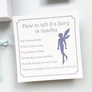K&ouml;nnte beinhalten: Eine quadratische wei&szlig;e Karte mit dekorativem Rand, die den violetten Text How to tell if a fairy is nearby und eine Liste von sechs Anzeichen wie sanfte Glockenkl&auml;nge und Feenstaub sowie eine violette Silhouette einer gefl&uuml;gelten Fee zeigt.