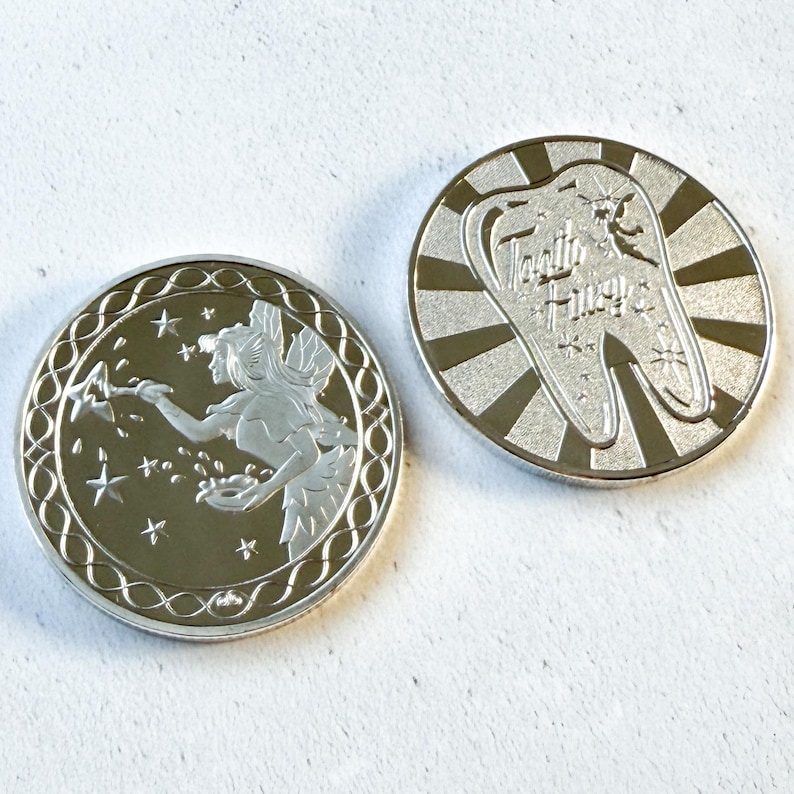 K&ouml;nnte beinhalten: Zwei kreisf&ouml;rmige silberfarbene Metallm&uuml;nzen. Eine M&uuml;nze zeigt eine gefl&uuml;gelte Fee mit einem Zauberstab, umgeben von Sternen. Die andere M&uuml;nze zeigt eine gro&szlig;e Zahngrafik mit dem Text Tooth Fairy und eine kleine Feensilhouette vor einem Strahlenhintergrund.
