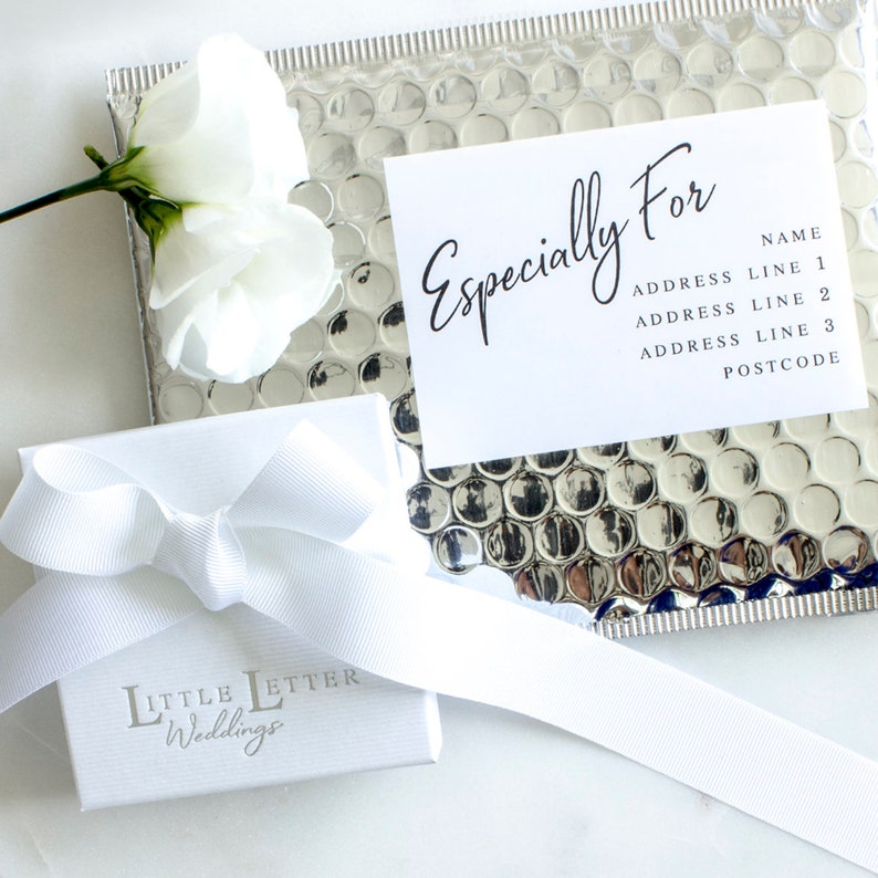 Op de afbeelding: Een witte cadeaudoos met een wit lint en de tekst "LITTLE LETTER Weddings" op de voorkant. Een witte kaart met de tekst "Especially For" en ruimte voor een adres ligt op een zilveren bubbeltjesenvelop.