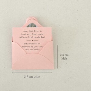 以下が含まれることがあります： 「every little letter is intricately hand made with no detail overlooked. little works of art delivered by your very own tooth fairy.」と書かれたメモが入った、淡いピンク色の小さな封筒。幅3.7cm、高さ2.5cmです。