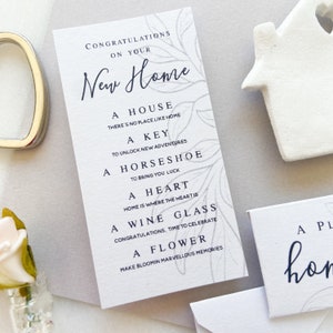 Op de afbeelding: Een witte kaart met een grijs bloemenpatroon en zwarte tekst die "Gefeliciteerd met je nieuwe huis" luidt. De tekst gaat verder met een lijst met items: "Een huis, Er is geen plek zoals thuis, Een sleutel, Om nieuwe avonturen te ontgrendelen, Een hoefijzer, Om geluk te brengen, Een hart, Thuis is waar het hart is, Een wijnglas, Gefeliciteerd, tijd om te vieren, Een bloem, Maak bloeiende, prachtige herinneringen."