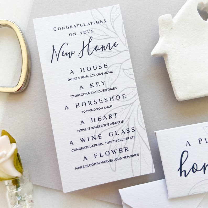Op de afbeelding: Een witte kaart met een grijs bloemenpatroon en zwarte tekst die "Gefeliciteerd met je nieuwe huis" luidt. De tekst gaat verder met een lijst met items: "Een huis, Er is geen plek zoals thuis, Een sleutel, Om nieuwe avonturen te ontgrendelen, Een hoefijzer, Om geluk te brengen, Een hart, Thuis is waar het hart is, Een wijnglas, Gefeliciteerd, tijd om te vieren, Een bloem, Maak bloeiende, prachtige herinneringen."