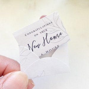 Op de afbeelding: Een witte kaart met een bloemenmotief en de tekst "Gefeliciteerd met je nieuwe huis Een huis" in zwarte letters.