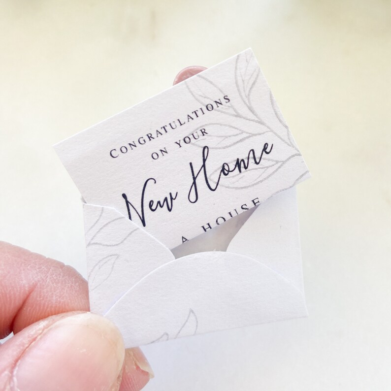 Op de afbeelding: Een witte kaart met een bloemenmotief en de tekst "Gefeliciteerd met je nieuwe huis Een huis" in zwarte letters.