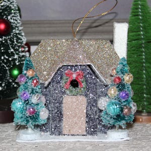 Puede incluir: Adorno navideño en miniatura con un techo dorado brillante y un exterior gris. La casa presenta una pequeña corona y está flanqueada por dos pequeños árboles de Navidad decorados. Se adjunta un hilo dorado para colgar.