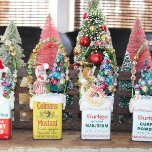 Peut inclure: Une décoration de Noël festive avec des boîtes d'épices recyclées ornées de mini-sapins, d'ornements et de personnages de vacances. Les boîtes portent les inscriptions "Schilling Whole Celery Seed", "Colmans Mustard", "Durkee Ground Marjoram" et "Durkee Curry Powder".
