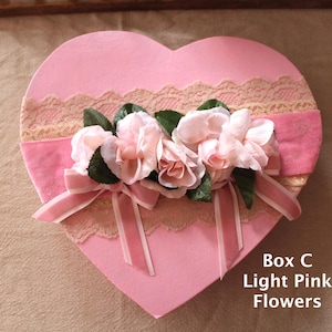 Puede incluir: Una caja en forma de corazón rosa claro decorada con flores rosa claro, hojas verdes y una cinta rosa. La caja tiene un ribete de encaje y el texto "Box C Light Pink Flowers".