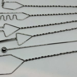May include: A set of six silver-coloured metal sculpting tools, each featuring a long, twisted handle and a unique wire-formed head. The shapes include a rectangle, a wavy line, a triangle, a pointed tip, a ball-end rod, and a pentagon.