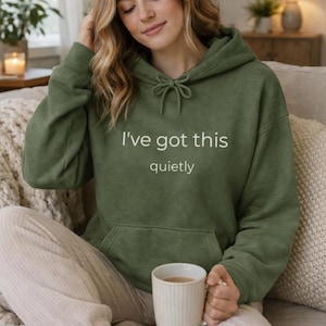 Op de afbeelding: Een olijfgroene hoodie met de tekst "I've got this quietly" in het wit. De hoodie heeft een trekkoord en een voorzak. De persoon houdt een witte mok met een drankje vast.