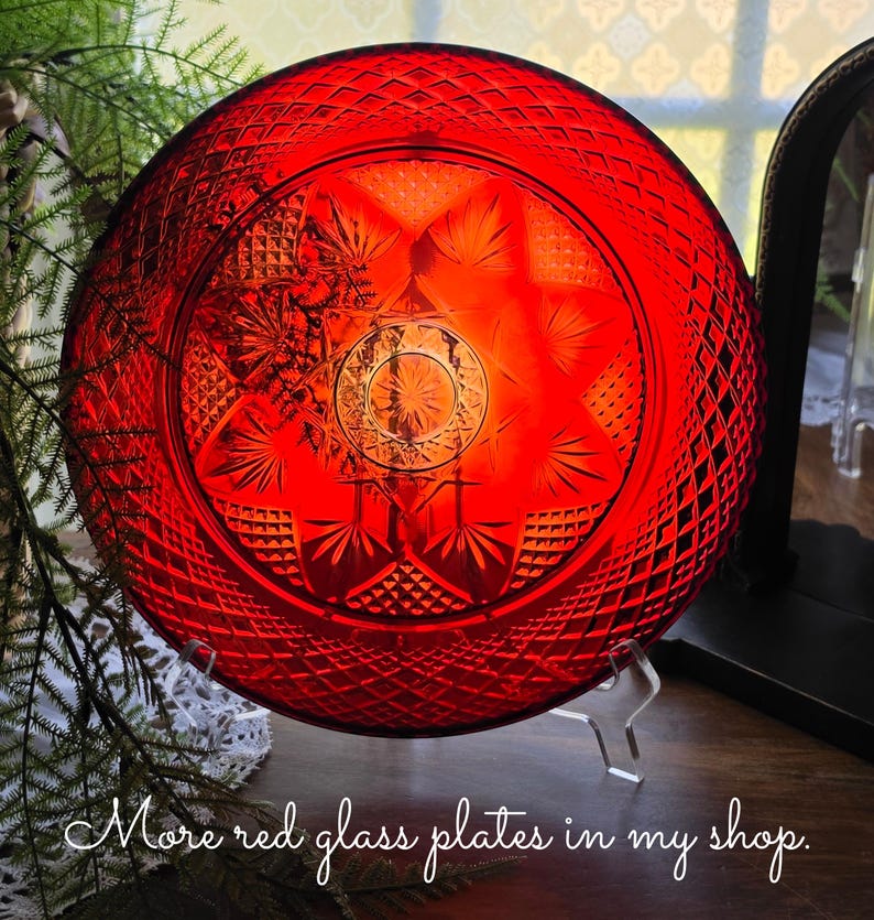 May include: A circular red glass plate featuring a pressed diamond pattern around the rim and a central starburst design with radiating leaf-like motifs. The plate is displayed on a clear stand and includes the text More red glass plates in my shop.
