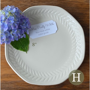 May include: An 8-inch off-white ceramic plate with a raised leaf pattern along the rim. A white tag reads Miss Purity White Your true-white color guide. A cluster of blue hydrangea flowers with green leaves rests on the left side of the plate.