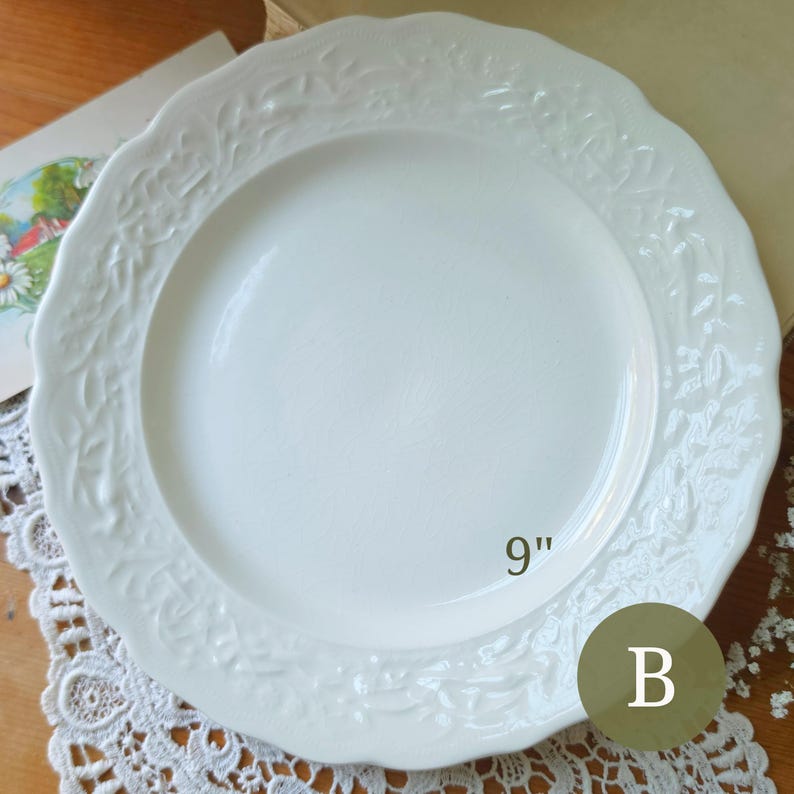 May include: A white ceramic dinner plate with a 9-inch diameter, featuring a smooth center and a wide, scalloped rim embossed with a detailed leaf and vine pattern. The plate is marked with the letter B in a circular overlay.