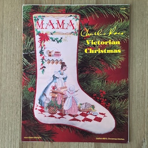May include: A cross-stitch pattern leaflet titled Charles Ross Victorian Christmas featuring a white stocking with a red border. The design shows a kitchen scene with the word MAMA, a person in a blue dress, a child, and a cat on a checkered floor.