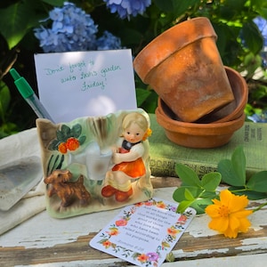 May include: A ceramic note holder featuring a child with a watering can and a small dog, holding a white card with the handwritten text 'Don't forget to water Leah's garden Friday!' and a green pen, alongside terracotta pots and a floral card.