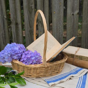 May include: A round, light-brown woven wicker basket with a single tall handle. Inside the basket sits an open book with cream-colored pages and text. A cluster of vibrant purple hydrangea flowers rests in the front of the basket.