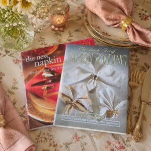 May include: Two hardcover books titled The New Napkin and The Simple Art of Napkin Folding by Linda Hetzer. The books feature illustrations of folded napkins with decorative rings, displayed on a floral tablecloth with gold cutlery and plates.