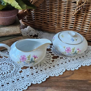 May include: A white porcelain creamer and sugar bowl set. The creamer has a handle and spout, while the sugar bowl has a lid. Both pieces are decorated with pink and blue floral patterns and gold trim. The set is on a white lace doily.