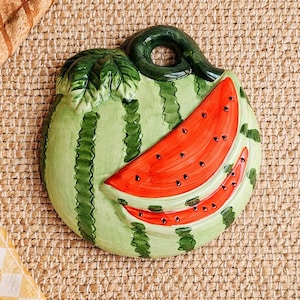 May include: A ceramic watermelon-shaped trivet. The trivet features a light green rind with dark green stripes and a red watermelon slice with black seeds. A green stem and leaf are attached to the top.