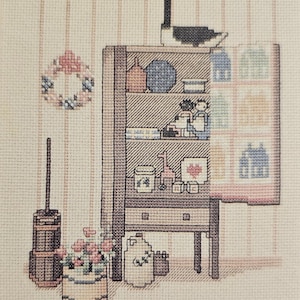 May include: Cross-stitch artwork depicting a cozy interior scene. A wooden cabinet is filled with various items, including a goose figurine, bottles, and small decorative objects. A floral wreath hangs on the wall, and a flower pot sits on the floor. The color palette is soft and pastel.