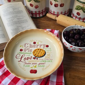 May include: A round ceramic pie dish with a scalloped edge, featuring a central graphic of a cherry pie and text that reads She's my Cherry Pie Homemade with Love Tastes sooo...good! along with illustrations of cherries and baking ingredients.