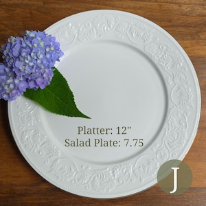 May include: A white round ceramic platter with an embossed leaf pattern along the rim, featuring a cluster of purple hydrangea flowers with a green leaf. Text on the platter reads: Platter: 12", Salad Plate: 7.75". A letter J is in a circle.