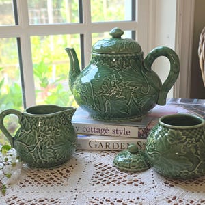 May include: A green ceramic teapot, creamer, and sugar bowl set featuring a raised relief pattern of grape leaves and vines. The teapot has a matching lid, and the sugar bowl has a separate lid. The pieces have a glossy finish and a checkered texture.